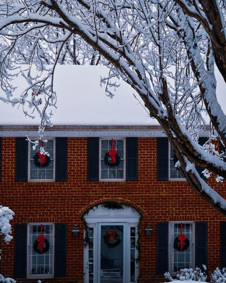 A brick colonial house in snow with a red wreath hung in every front window