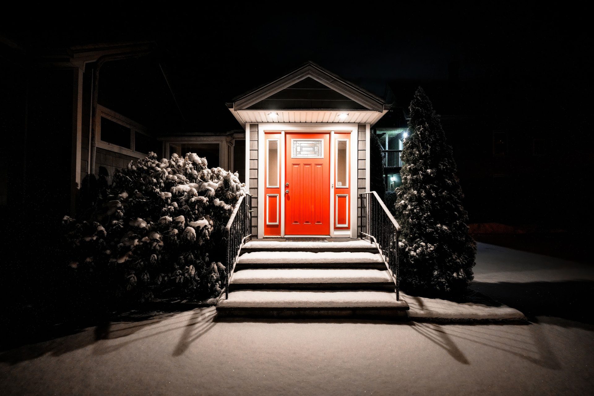 A red front door at the top of lit steps on a winter night, with clipped evergreens either side and snow on the ground