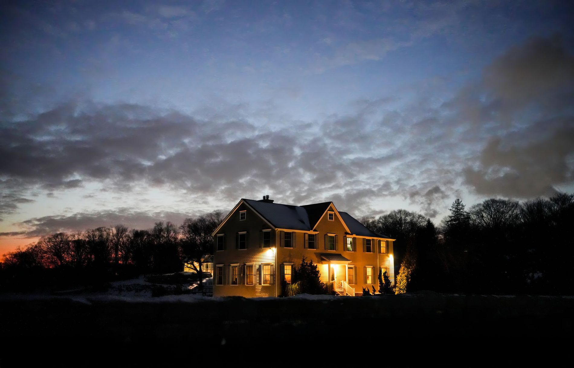 A two story house lit warm along the roofline against a heavy dusk sky