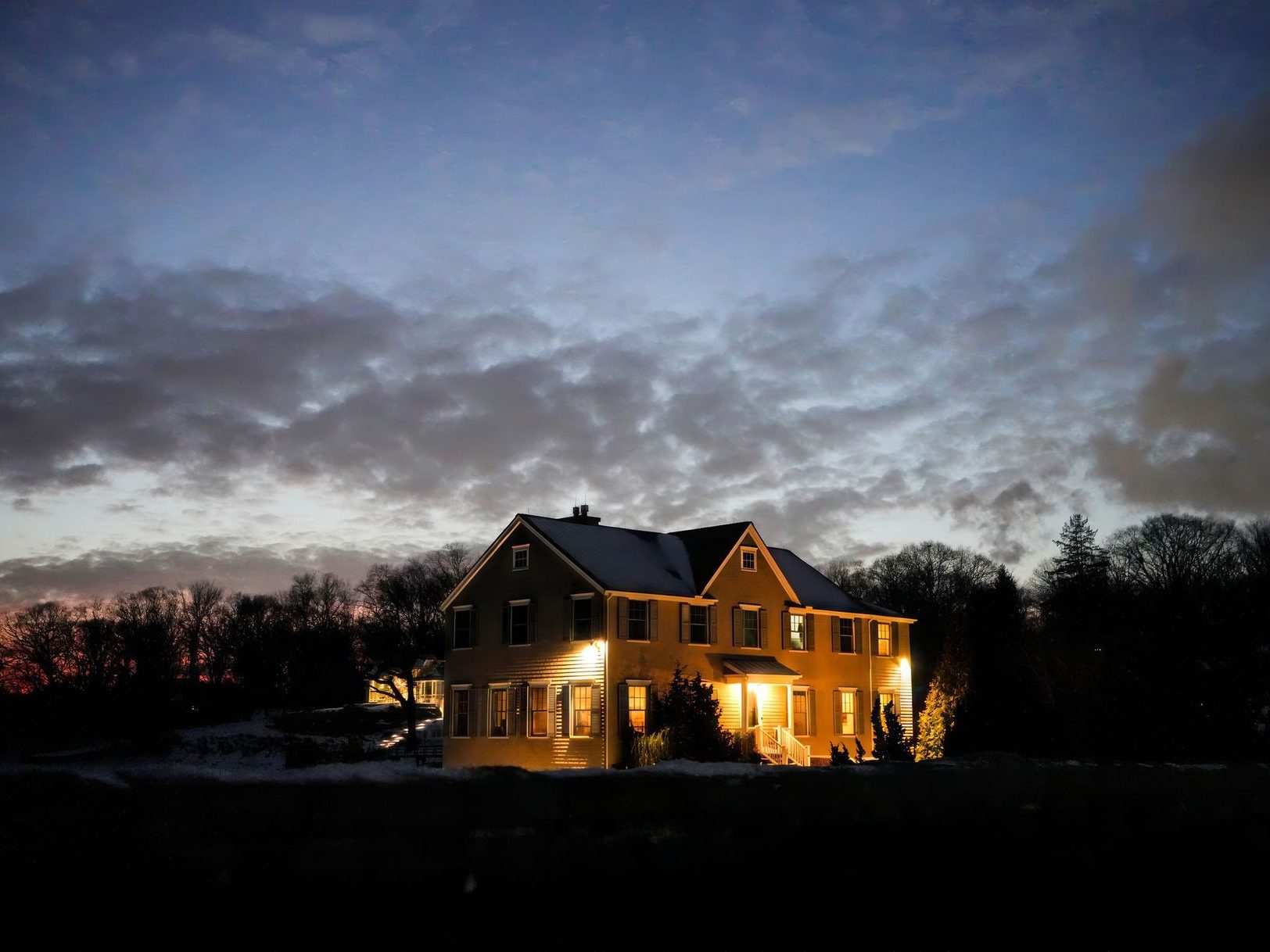 A two story house lit warm against a heavy dusk sky