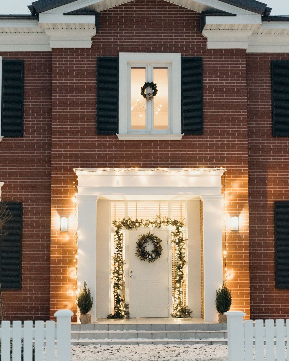 A white portico outlined in warm white bulbs with garland on the columns and snow across the lawn