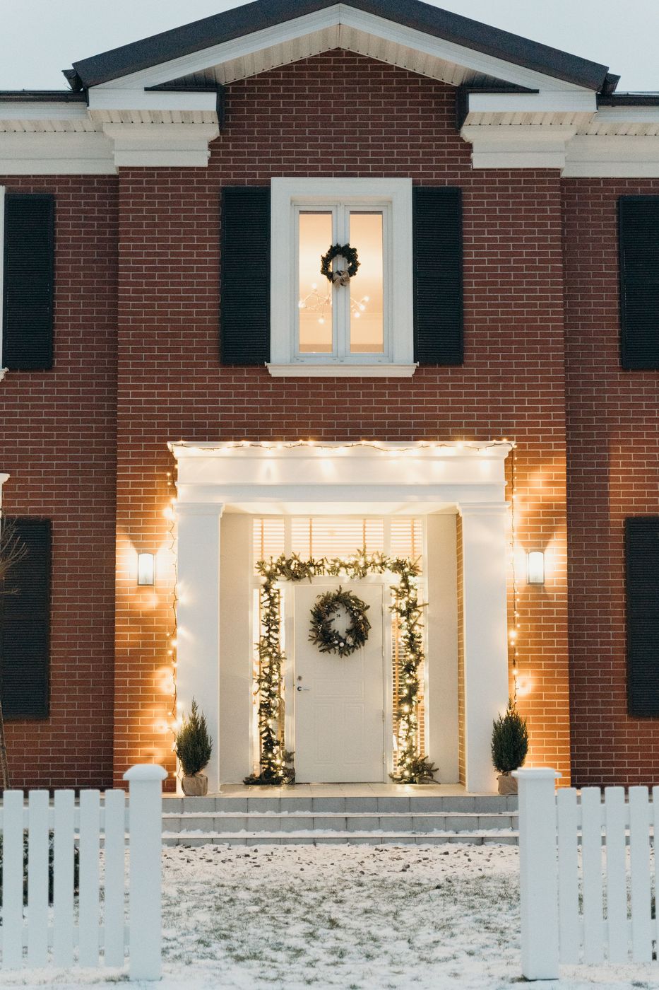 Warm white bulbs outlining a white portico with garland on the columns and snow on the lawn